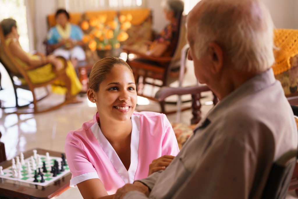 Old people in geriatric hospice: young attractive hispanic woman working as nurse takes care of a senior man on wheelchair. She talks with him then goes away to help other patients